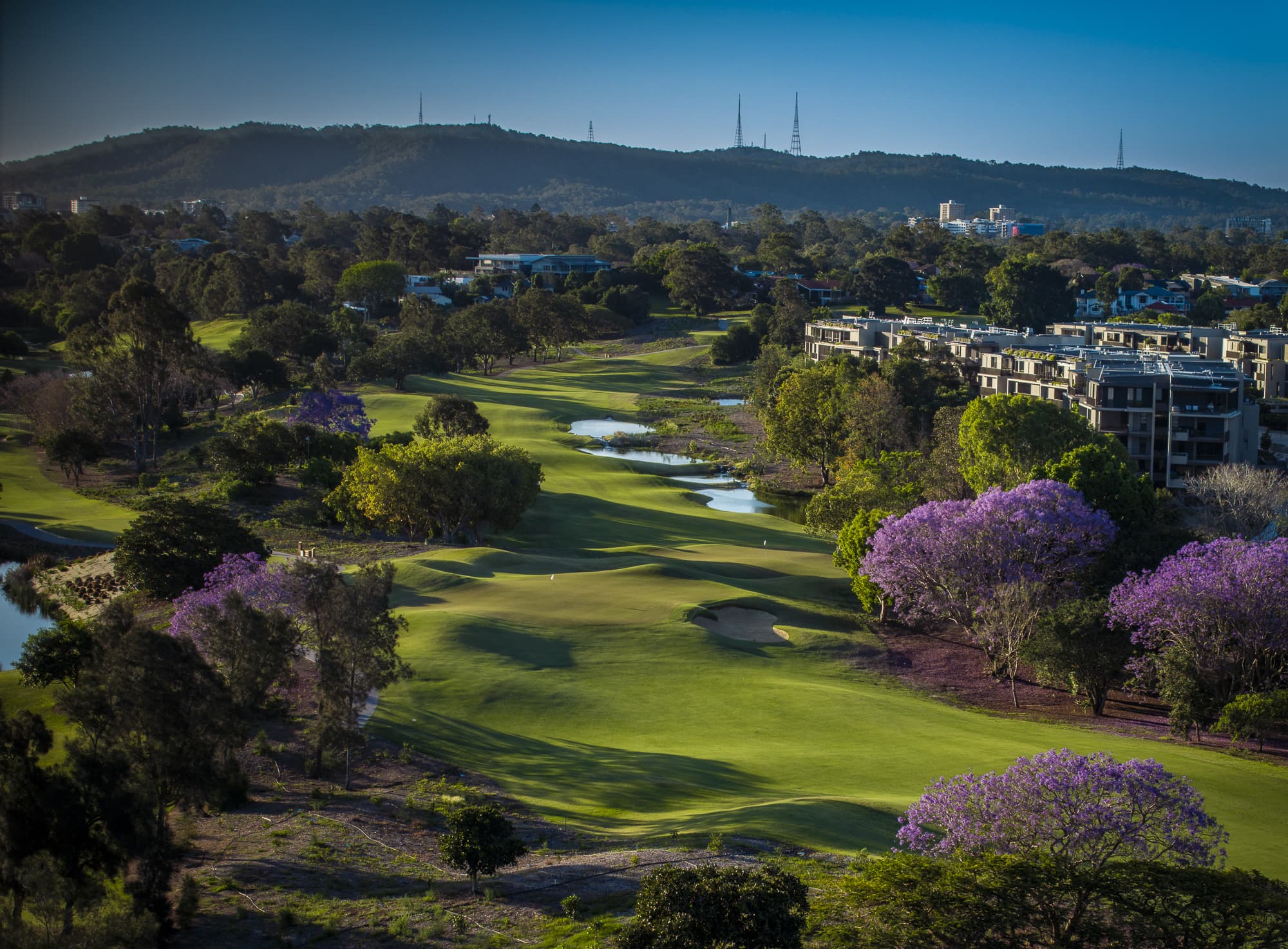 A scenic view of a golf course surrounded by lush greenery and purple-flowering trees. Buildings are visible to the right, with hills and a clear blue sky in the background.